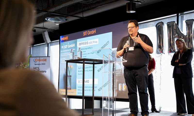 A man speaks into a microphone on stage at a conference, standing in front of a presentation screen. A woman stands nearby and an audience member is visible in the foreground. 